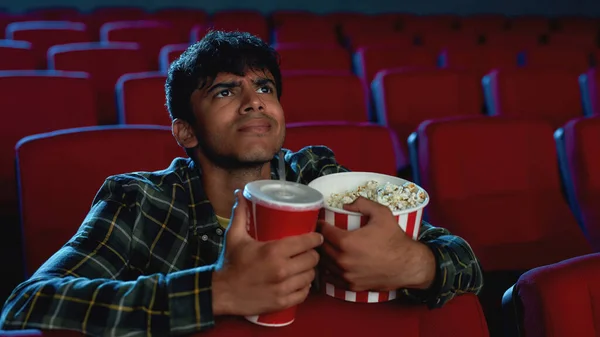 Portrait of concentrated young guy holding a drink and popcorn basket while watching movie alone in empty theater auditorium — Stock Photo, Image