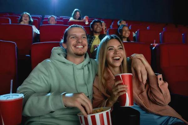 Porträt eines fröhlichen jungen Paares, das lacht, Getränke und Popcorn trinkt, während es gemeinsam im Kinosaal sitzt — Stockfoto