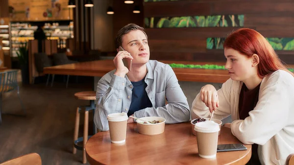 Twee tieners samen, zitten overdag in een café. Meisje kijkt verveeld, eet haar maaltijd terwijl haar vriend haar negeert, praat aan de telefoon — Stockfoto