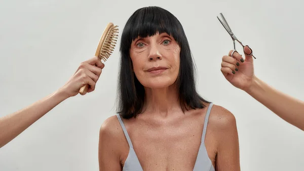 Duas mãos com escova de cabelo e tesoura fazendo corte de cabelo para uma mulher semi-nua sênior, isolado sobre fundo cinza — Fotografia de Stock