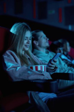 Güzel genç bayan gülümsüyor, sinemadaki sıkıcı filmi kaçırırken telefonunu kullanıyor.