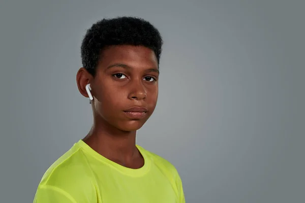 Ouvir música durante o treino. Retrato de um adolescente africano usando fones de ouvido sem fio olhando para a câmera enquanto estava de pé contra fundo cinza em estúdio — Fotografia de Stock