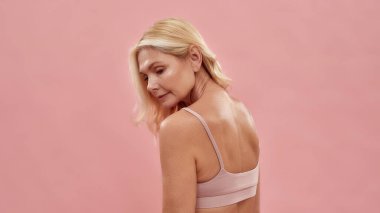 Doğal güzellik. Güzel, orta yaşlı, beyaz, sarışın bir kadının, pembe arka planda yarı çıplak poz verişi.