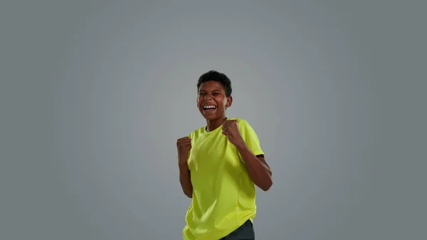 Eccitato felice ragazzo africano adolescente indossa vestiti sportivi tenendo le mani in pugno mentre celebra il successo, godendo la vittoria, in piedi su sfondo grigio — Foto Stock