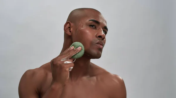 Portret przystojnego afrykańskiego młodego mężczyzny korzystającego z zabiegu pielęgnacji skóry, używającego gąbki oczyszczającej podczas mycia twarzy na szarym tle — Zdjęcie stockowe