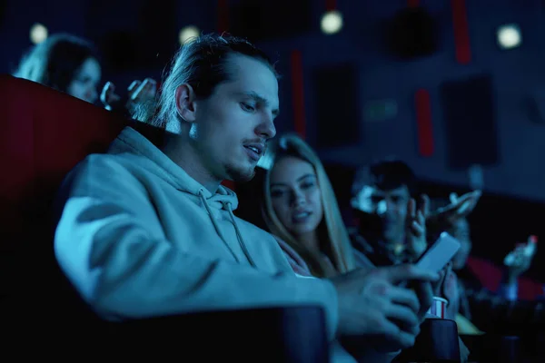 映画館で映画のデートをしながら、彼のスマートフォンを使用して若い男に焦点を当て — ストック写真