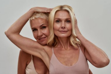 Stüdyo portresi iki güzel sarışın kadın kameraya bakıyor, yan yana duruyorlar, gri arka planda yalnız poz veriyorlar.