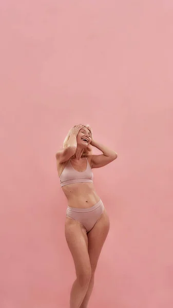 Feeling good. Playful middle aged caucasian blonde woman wearing underwear keeping eyes closed and smiling while posing isolated over pink background — стоковое фото