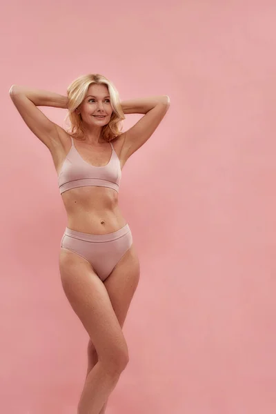 Feeling beautiful at any age. Happy attractive middle aged caucasian woman in underwear smiling and looking away while standing against pink background, vertical shot