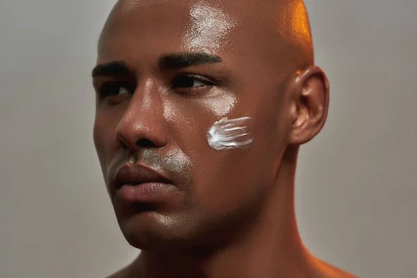 Retrato de close-up de belo jovem afro-americano com creme aplicado em sua bochecha olhando para o lado enquanto posando isolado sobre fundo cinza — Fotografia de Stock