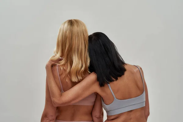 Rear view of two mature caucasian women in underwear standing together while posing isolated over light background