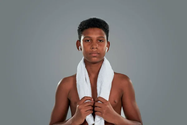 Po tréninku. Polonahý dospívající africký chlapec s ručníkem na ramenou dívá do kamery, stojí proti šedému pozadí, dělá sportovní cvičení — Stock fotografie