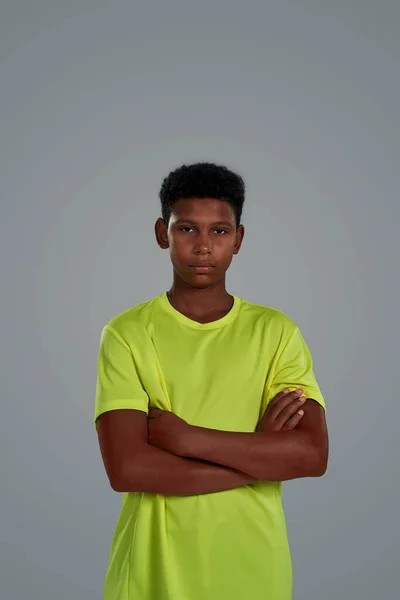 Tiro vertical de niño africano adolescente deportivo confiado con camiseta de neón manteniendo los brazos cruzados mientras posa aislado sobre un fondo gris — Foto de Stock