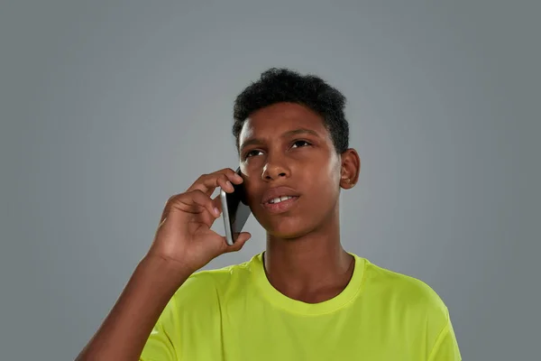 Retrato de um adolescente africano vestindo camiseta de néon falando por telefone enquanto estava de pé contra fundo cinza, tiro vertical — Fotografia de Stock