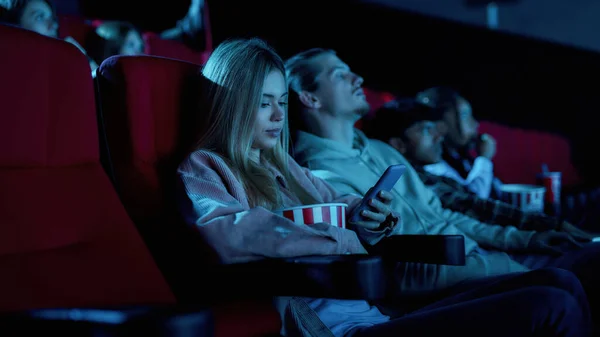 Mooie jonge vrouw met behulp van haar telefoon voor het sms 'en terwijl het missen van saaie film in de bioscoop — Stockfoto
