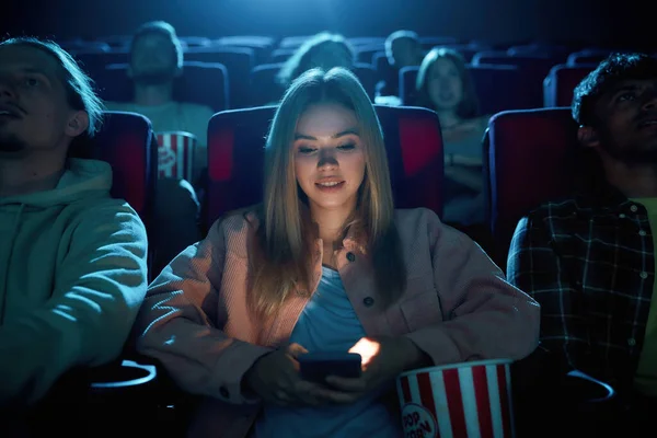 Schöne junge Frau lächelt und benutzt ihr Handy für SMS, während sie einen Film im Kino anschaut — Stockfoto