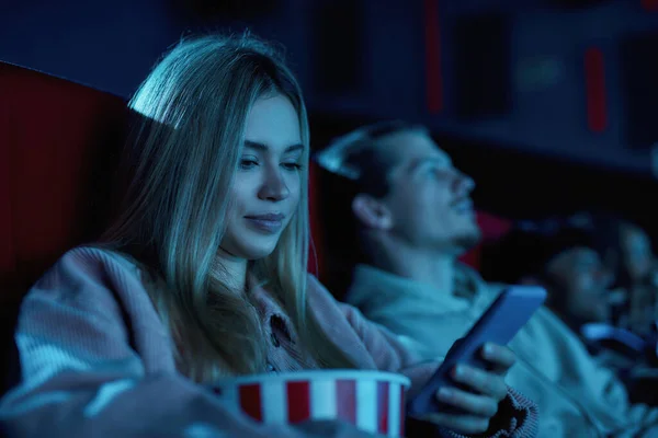 Portret van een mooie jonge vrouw glimlachend, met behulp van haar telefoon terwijl het missen van saaie film in de bioscoop — Stockfoto