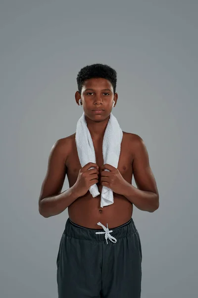 Resting after workout. Half naked teenage african boy with towel on shoulders wearing wireless earphones looking at camera while standing against grey background — Stock Photo, Image