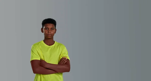 Sporty teenage african boy wearing neon t shirt keeping arms crossed while posing isolated over grey background in studio, side view — Stock Photo, Image