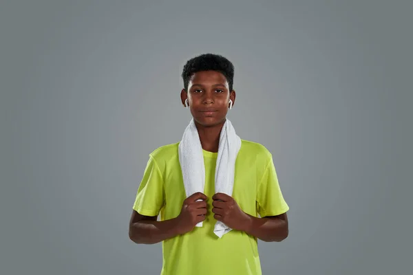 Retrato de um menino africano adolescente feliz em camisa de néon com toalha branca nos ombros usando fones de ouvido sem fio olhando para a câmera enquanto posando contra fundo cinza — Fotografia de Stock