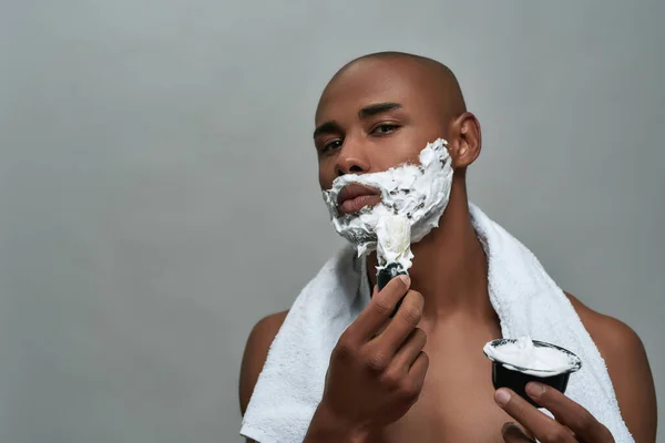 Pemuda african Amerika yang tidak menarik melihat kamera, menggunakan kuas sambil mengenakan busa cukur di wajahnya, berpose terisolasi di atas latar belakang abu-abu — Stok Foto