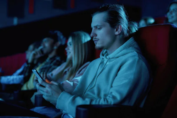 Konzentrierter junger Mann benutzt sein Smartphone, während er einen langweiligen Film im Kino verpasst — Stockfoto