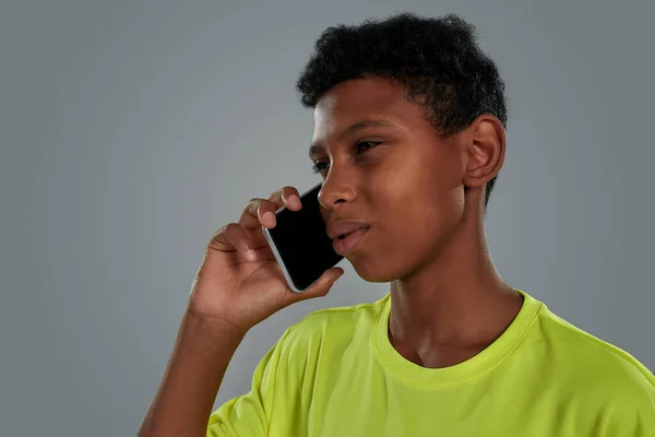 Foto ravvicinata di un ragazzo africano adolescente che indossa una maglietta al neon mentre parla per telefono mentre si trova su uno sfondo grigio — Foto Stock