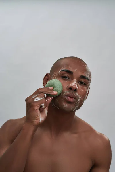 Портрет привлекательного африканского молодого человека, использующего очищающую губку во время мытья лица, стоящего изолированно на сером фоне — стоковое фото