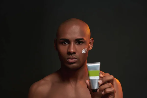 Portrait of shirtless african american man looking at camera, holding beauty product while posing with cream applied on his cheek isolated over black background — Stock Photo, Image