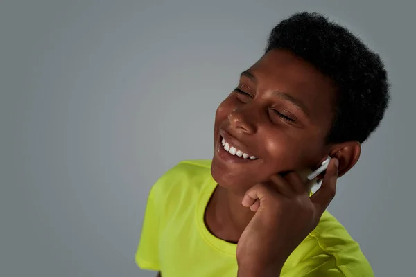 Menino africano adolescente feliz usando fones de ouvido sem fio ouvindo música durante o treino, mantendo os olhos fechados enquanto estava de pé contra fundo cinza — Fotografia de Stock