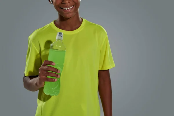 Tiro recortado de um menino africano adolescente vestindo camiseta de néon segurando bebida energética e sorrindo para a câmera enquanto estava de pé contra fundo cinza — Fotografia de Stock