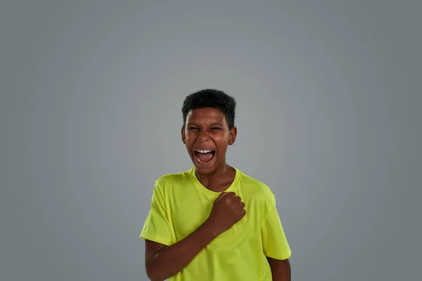 Портрет мотивованого і схвильованого підлітка африканського хлопчика в сорочці неону, що тримає кулак на грудях і кричить, стоячи на сірому фоні — стокове фото
