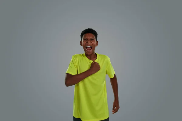 Ich bin ein Gewinner. Motivierter und aufgeregter afrikanischer Teenager in neonfarbenem T-Shirt, die Faust auf der Brust und schreiend vor grauem Hintergrund — Stockfoto