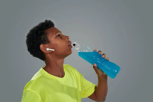 Вид сбоку на африканского мальчика в беспроводных наушниках, пьющего синий энергетический напиток, стоя на сером фоне — стоковое фото
