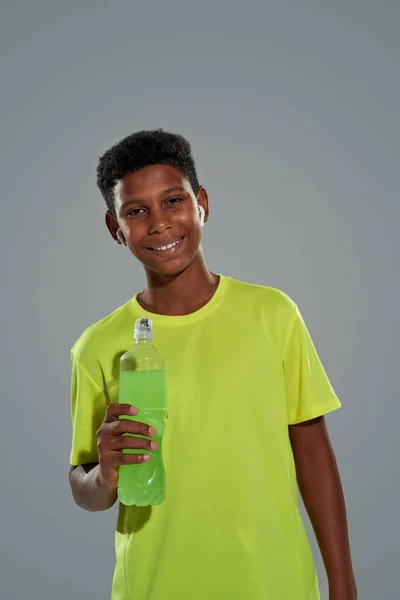 Вертикальний знімок щасливого підлітка африканського хлопчика в спортивному одязі, що тримає енергетичний напій і посміхається на камеру, стоячи на сірому фоні — стокове фото