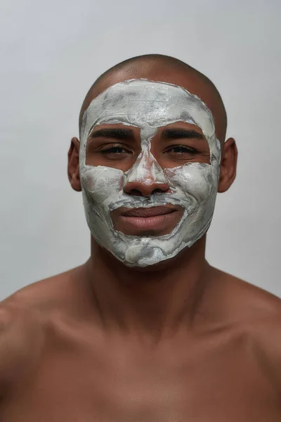 Close-up portret van vrolijke jonge Afrikaan amerikaanse man knipogen op camera, met behulp van gezicht klei masker, poseren geïsoleerd over grijze achtergrond — Stockfoto
