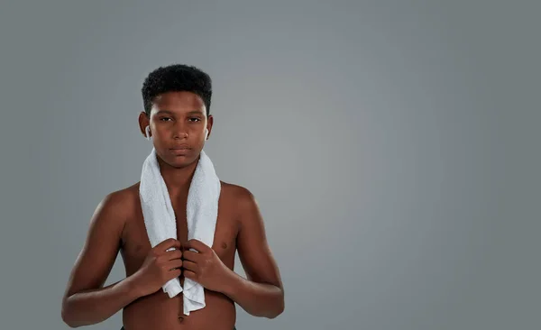 Medio desnudo adolescente africano chico con toalla en los hombros con auriculares inalámbricos mirando a la cámara mientras está de pie sobre fondo gris — Foto de Stock