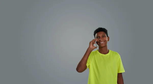 Joyeux adolescent africain portant un t-shirt néon parlant par téléphone et souriant à la caméra tout en se tenant debout sur fond gris, espace de copie pour le texte — Photo