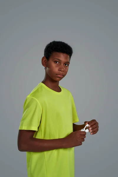 Menino africano adolescente tirando um fone de ouvido sem fio enquanto estava de pé contra fundo cinza em estúdio, olhando para a câmera — Fotografia de Stock
