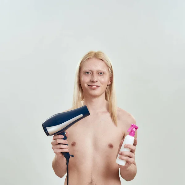 Young blonde man holding hair dryer and fixation liquid — Stock Photo, Image