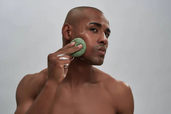 Portrait of handsome african american young man enjoying skincare procedure, using cleansing sponge while washing his face isolated over gray background — Stock Photo, Image