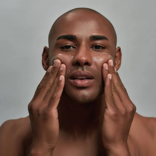 Retrato de primer plano del guapo joven afroamericano mirando a la cámara, aplicándose crema en la cara, posando aislado sobre un fondo gris — Foto de Stock