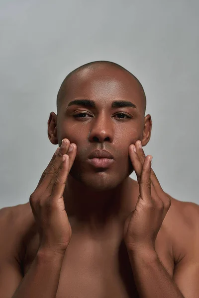 Póló nélküli, fiatal afro-amerikai férfi portréja, amint a kamerába néz, miközben testápolót használ az arcára, szürke háttérrel elszigetelve pózol. — Stock Fotó