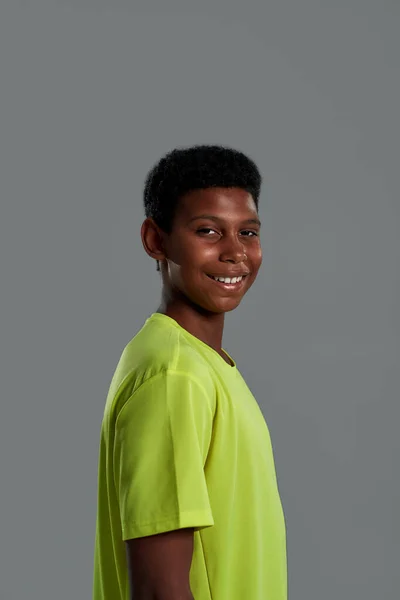 Vista lateral de um adolescente feliz e alegre menino africano sorrindo para a câmera enquanto posando isolado sobre fundo cinza no estúdio — Fotografia de Stock