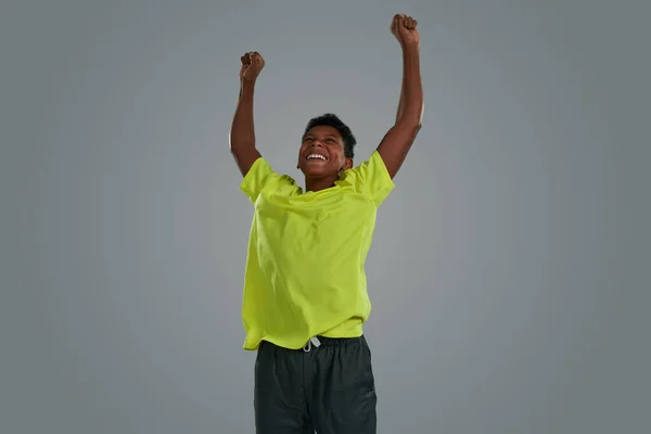 Winnen. opgewonden tiener afrikaanse jongen dragen neon t shirt vieren succes, houden armen omhoog terwijl staande tegen grijze achtergrond — Stockfoto