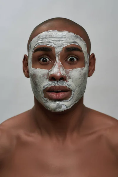 Közelkép portré meglepett fiatal afro-amerikai férfi néz kamera, az arc agyag maszk, pózol elszigetelt szürke háttér — Stock Fotó