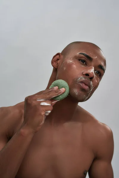 Portrait of handsome african american young man using cleansing sponge for washing face, standing isolated over gray background — Stock Photo, Image