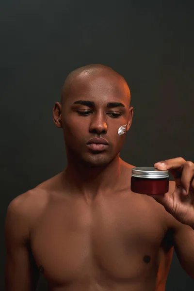 Shirtless Afrikaanse Amerikaanse man holding, kijken naar schoonheid product terwijl poseren met crème aangebracht op zijn wang geïsoleerd over zwarte achtergrond — Stockfoto