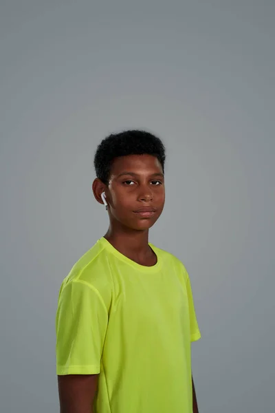 Vertical shot of a teenage african boy wearing wireless earphones looking at camera, enjoying listening music while standing against grey background — Stock Photo, Image