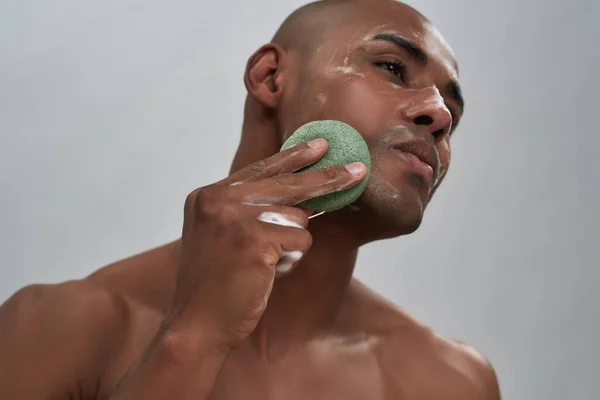 Close up portrait of attractive african american young man enjoying skincare procedure, using cleansing sponge while washing his face isolated over gray background — Stock Photo, Image